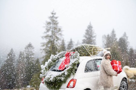 Téléchargez les photos : Jeune femme avec un coffret cadeau près de sa voiture avec arbre de Noël sur un toit lors d'une chute de neige, voyageant en véhicule très à la montagne pendant les vacances d'hiver - en image libre de droit