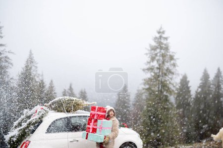 Foto de La mujer sostiene un montón de cajas de regalo mientras está de pie junto al coche decorado con una corona de Navidad y un árbol en una azotea, viajando en coche por las montañas con clima nevado. Concepto de vacaciones de invierno - Imagen libre de derechos