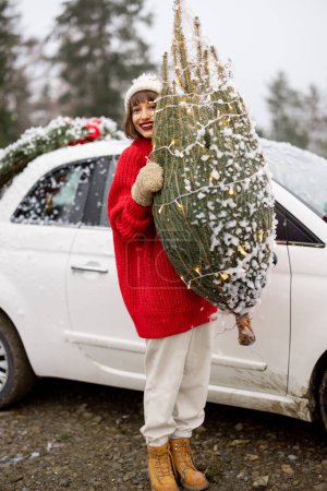 Téléchargez les photos : Portrait d'une femme avec arbre de Noël enveloppé, voyageant en voiture dans les montagnes par temps neigeux. Concept de préparation pour des vacances d'hiver heureuses. Véhicule décoré avec des cadeaux et une couronne derrière - en image libre de droit