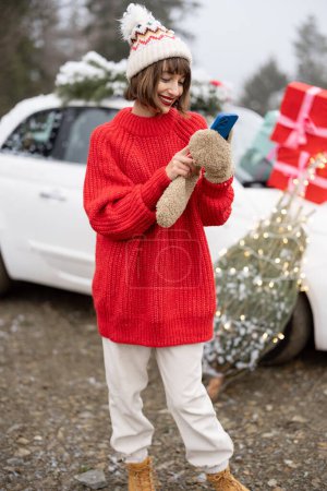 Téléchargez les photos : Femme en pull rouge en utilisant le téléphone tout en se tenant avec l'arbre de Noël et présente près de la voiture dans les montagnes. Concept de préparation pour des vacances d'hiver heureuses - en image libre de droit