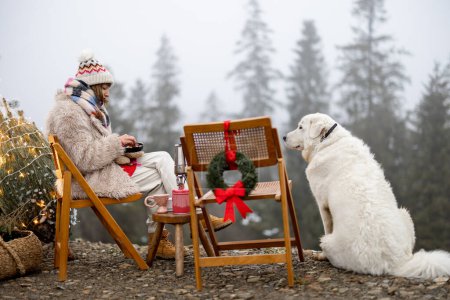 Foto de Mujer joven se relaja y disfruta de la calma en la naturaleza, mientras se sienta con su perro en el picnic durante las vacaciones de invierno en las montañas. Celebrando el Año Nuevo mientras viaja en coche con mascota - Imagen libre de derechos