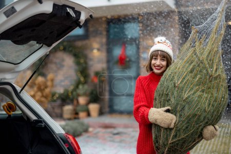 Téléchargez les photos : Portrait of young cheerful woman in red sweater and hat holding wrapped Christmas on porch of her house decorated for winter holidays. Concept of preparing and decorations for New Years holidays - en image libre de droit
