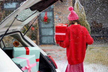 Foto de Mujer en suéter rojo y sombrero lleva envuelto el árbol de Navidad y un regalo a su casa en la caída de nieve, Vista desde atrás. Concepto de preparación para las vacaciones de invierno - Imagen libre de derechos