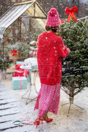 Photo for Young woman in red decorates lush Christmas tree with festive bows at backyard of her house on snow fall, preparing for a winter holidays - Royalty Free Image