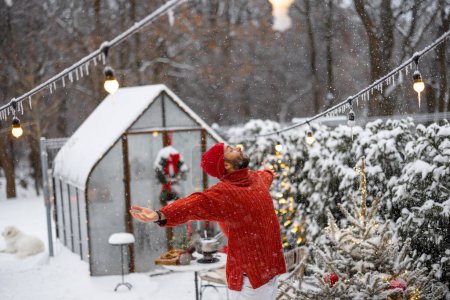 Foto de El hombre de rojo levanta las manos sintiéndose libre y feliz durante el invierno en el patio trasero cubierto de nieve. Concepto de felices vacaciones de invierno - Imagen libre de derechos