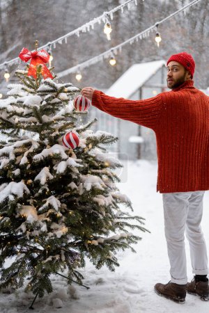 Téléchargez les photos : Mann decorates Christmas tree with festive ballls while stands on terrace at snowy backyard. Girl preparing for a winter holidays - en image libre de droit