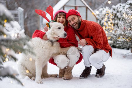Foto de Lovely couple hug with their cute dog wearing toy deers horns at snowy backyard. Young family spending happy winter time together outdoors - Imagen libre de derechos