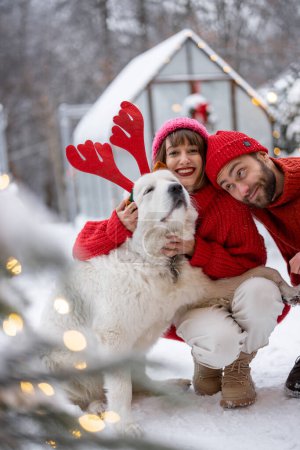 Photo for Lovely couple hug with their cute dog wearing toy deers horns at snowy backyard. Young family spending happy winter time together outdoors - Royalty Free Image