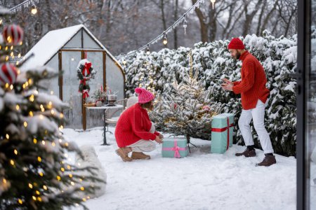 Téléchargez les photos : Man and woman put presents under Christmas tree while decorating backyard for a winter holidays. Happy family celebrating New Years holidays outdoors - en image libre de droit