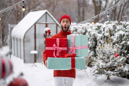 Foto de Cheerful man in red sweater and hat carries gift boxes at snowy backyard. Concept of happy winter time, Thanks giving day and Valentine holiday - Imagen libre de derechos