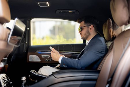 Photo for Businessman sits with phone on a backseat of luxury car. Man doing business on the road in prestigious car. Concept of business transportation - Royalty Free Image