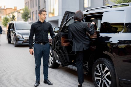 Téléchargez les photos : Homme chauffeur aide une femme d'affaires à monter dans une voiture, ouvrant la porte d'un taxi SUV de luxe. Femme d'affaires avec sac à main portant une tenue formelle noire. Concept de service de transport - en image libre de droit
