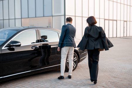Téléchargez les photos : Homme d'affaires et femme d'affaires marchent ensemble à la voiture sur le parking près du bâtiment moderne, va conduire en voiture de luxe. L'homme ouvre la porte du véhicule - en image libre de droit