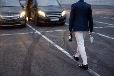 Foto de Empresario camina con bebida alcohólica a los coches al aire libre, teniendo una celebración informal con los socios en el estacionamiento - Imagen libre de derechos