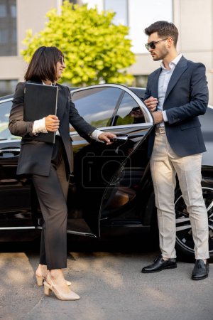 Téléchargez les photos : Une conductrice aide un homme d'affaires à sortir de la voiture. ouvrir une porte. Concept de chauffeur personnel, taxi de luxe ou voyages d'affaires - en image libre de droit
