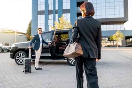 Foto de Elegante mujer de negocios camina hacia el coche de lujo, hombre con una maleta abre la puerta del vehículo, dejando entrar a la dama. Concepto de viaje de negocios y viajes - Imagen libre de derechos