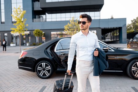 Téléchargez les photos : Élégant homme d'affaires marche avec une valise, portant une veste devant la voiture près du bâtiment moderne au coucher du soleil - en image libre de droit
