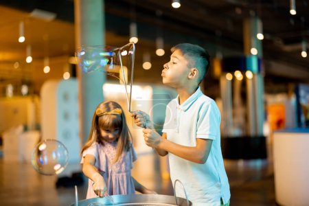 Téléchargez les photos : Petite fille et garçon font des bulles de savon, tout en jouant ensemble et en s'amusant dans un musée des sciences. Concept de divertissement et d'apprentissage pour enfants - en image libre de droit