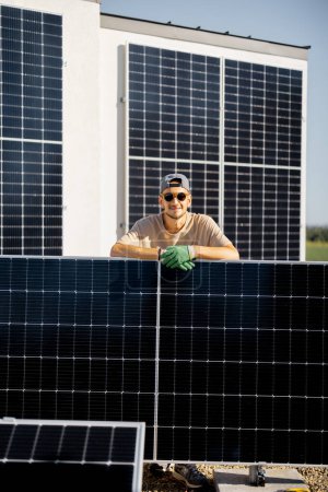 Téléchargez les photos : Portrait d'un homme debout avec un panneau solaire sur un toit de sa maison pendant le processus d'installation. Propriétaire de la propriété installant des panneaux solaires pour l'autoconsommation - en image libre de droit