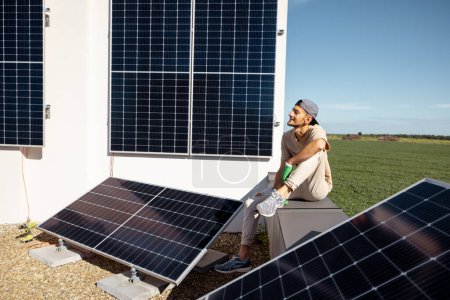 Téléchargez les photos : Un homme assis sur un toit de sa maison avec une centrale solaire installée dessus. Concept d'énergie verte renouvelable et de durabilité - en image libre de droit