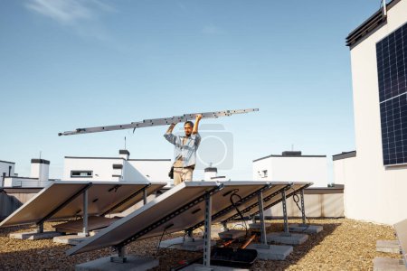 Téléchargez les photos : L'homme porte une échelle lors de l'installation de panneaux solaires sur le toit d'une maison privée par une journée ensoleillée. Vue grand angle d'en bas. Concept d'entretien et d'installation d'une centrale solaire - en image libre de droit