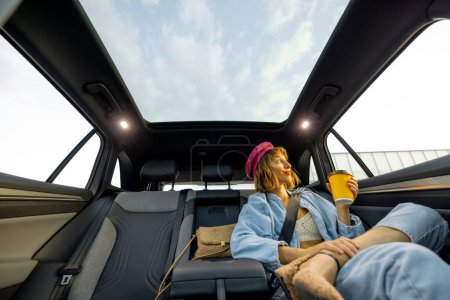 Photo for Stylish woman travels sits relaxed with coffee cup on backseat of a car. Wide angle view, modern car with panoramic rooftop - Royalty Free Image