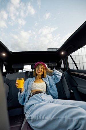 Foto de Mujer joven y elegante disfruta de la música mientras viaja en coche, sentado relajado con una taza de café en el asiento trasero. Amplia vista angular, coche moderno con techo panorámico - Imagen libre de derechos