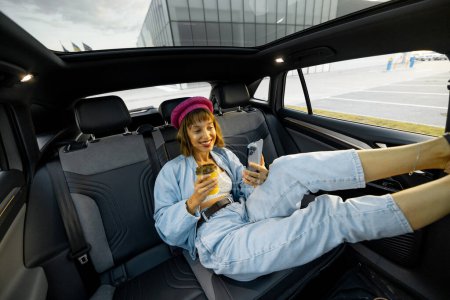 Foto de Joven mujer con estilo disfruta de viajar en coche, sentado relajado con una taza de café y teléfono en el asiento trasero. Amplia vista angular, coche moderno con techo panorámico - Imagen libre de derechos