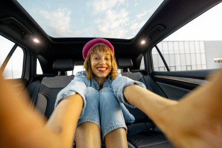 Téléchargez les photos : Jeune femme élégante fait un selfie tout en étant assis sur le siège arrière d'une voiture. Dame voyageant joyeusement en véhicule moderne avec toit panoramique - en image libre de droit