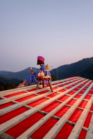 Foto de La mujer disfruta de una hermosa puesta de sol en las montañas, mientras está sentada en el techo de la casa inacabada. Concepto de romance, casa de ensueño y escapada a la naturaleza - Imagen libre de derechos