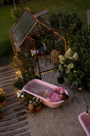 Téléchargez les photos : Cour arrière confortable avec une femme se baignant dans une baignoire rose à l'extérieur au crépuscule. Concept de détente et de beauté - en image libre de droit