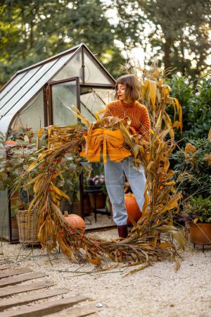 Téléchargez les photos : Portrait d'une femme avec une grande couronne de maïs, décorant son jardin pour des vacances d'automne. Concept de décoration, vacances de Thanksgiving et jardinage - en image libre de droit