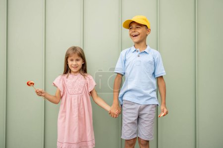 Téléchargez les photos : Portrait d'un enfant heureux avec des bonbons sucrés sur fond de mur vert à l'extérieur, passer l'été ensemble. Petit garçon avec une fille visitant le parc d'attractions et s'amusant - en image libre de droit