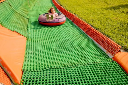 Foto de Niña feliz desciende en tubo de trineo inflable en una pista de verano, divirtiéndose en el parque de atracciones. Concepto de vacaciones de verano y entretenimiento infantil - Imagen libre de derechos
