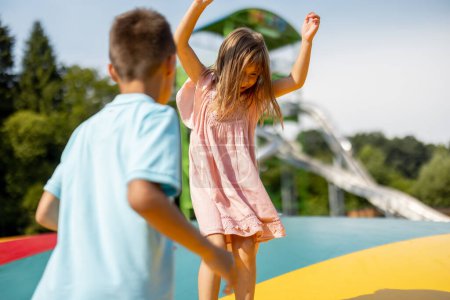 Téléchargez les photos : Les enfants sautant sur le trampoline gonflable, s'amusant visitant le parc d'attractions pendant des vacances d'été. Frère avec une sœur passer du temps libre ensemble - en image libre de droit