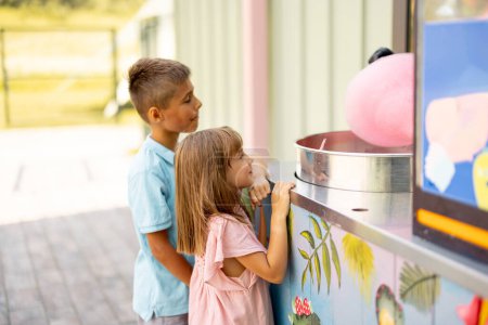 Foto de Niña y niño esperando que un dulce algodón de azúcar se haga en el mostrador mientras visita el parque de atracciones durante unas vacaciones de verano - Imagen libre de derechos