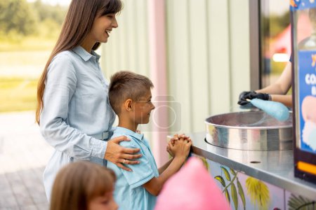 Photo for Mom with kids waiting for a sweet cotton candy to be made at the counter shop while visiting amusement park during a summer vacation - Royalty Free Image