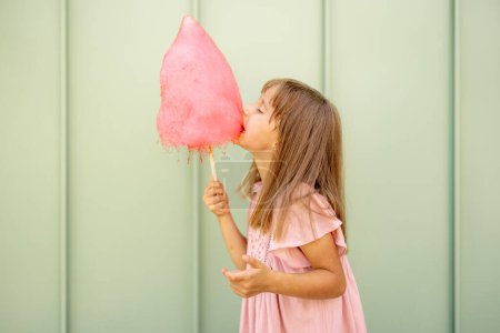 Photo for Portrait of a little girl with pink cotton candy on green wall background outdoors. Happy kids visiting amusement park and having fun - Royalty Free Image