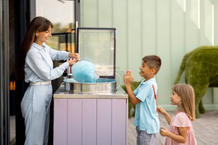 Foto de Niña y niño esperando un dulce algodón de azúcar para ser hecho por el vendedor femenino en la tienda al aire libre del parque de atracciones durante unas vacaciones de verano - Imagen libre de derechos