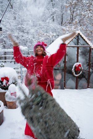 Foto de Mujer de rojo disfruta de las vacaciones de invierno de pie con el árbol de Navidad y lanzando feliz nieve en el patio trasero - Imagen libre de derechos
