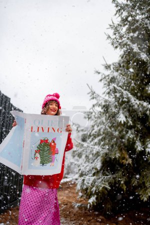 Téléchargez les photos : Femme lit le magazine Nouvel An tout en se tenant à l'extérieur devant le sapin neigeux. Concept de vacances d'hiver et humeur - en image libre de droit