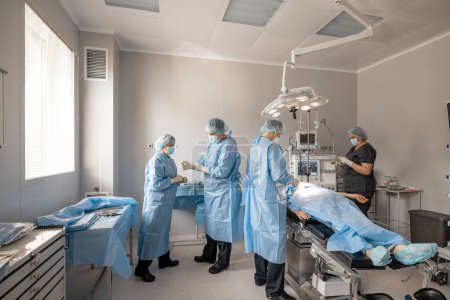 Foto de Enfermera con cirujano preparándose para una operación, vistiendo uniforme estéril en quirófano con un paciente acostado. Concepto de esterilidad durante el tratamiento quirúrgico - Imagen libre de derechos