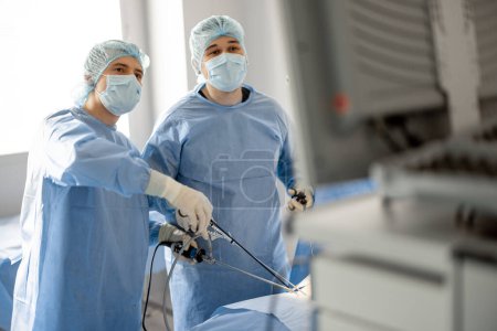 Téléchargez les photos : Deux chirurgiens en uniforme opèrent un patient avec des endscopes et regardent dans un moniteur pendant une chirurgie invasive minimale. Concept de traitement endoscopique assisté par ordinateur - en image libre de droit