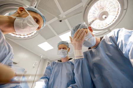 Foto de Los cirujanos miran el lugar de la intervención quirúrgica con horror y desesperación durante una operación, vista desde abajo. Descubrimiento de lo terrible durante la disección quirúrgica - Imagen libre de derechos