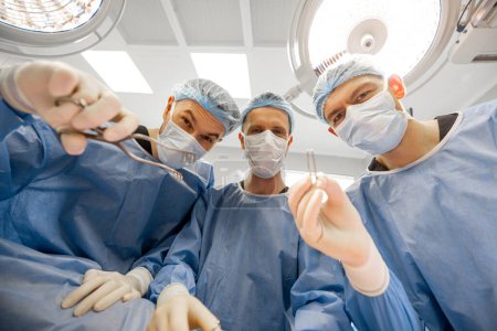 Foto de Retrato de tres cirujanos aterradores con herramientas quirúrgicas que miran a la cámara en el quirófano - Imagen libre de derechos
