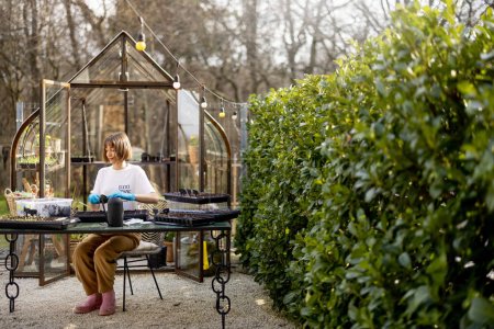Téléchargez les photos : Une jeune femme sème des graines dans des plateaux alors qu'elle est assise à table dans son beau jardin au début du printemps. Concept de passe-temps et mode de vie écologique durable - en image libre de droit