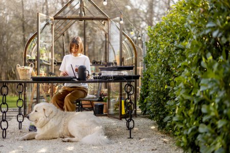 Photo for Woman planting flower seeds into seedling trays while sitting by the table with her dog outdoors. Concept of a hobby or active leisure time in garden - Royalty Free Image