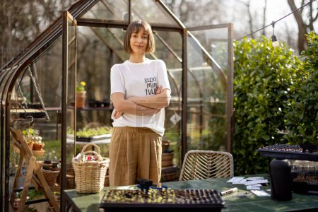 Téléchargez les photos : Portrait d'une jeune jardinière debout dans un beau jardin au début du printemps avec une serre derrière - en image libre de droit