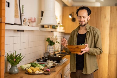 Foto de Hombre guapo mezclando ensalada en un tazón de madera mientras cocina comida vegetariana saludable en el elegante interior de la cocina casera - Imagen libre de derechos