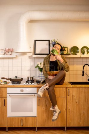 Téléchargez les photos : Portrait d'un homme avec des feuilles de salade assis sur le dessus de la table tout en cuisinant des aliments sains à la maison - en image libre de droit